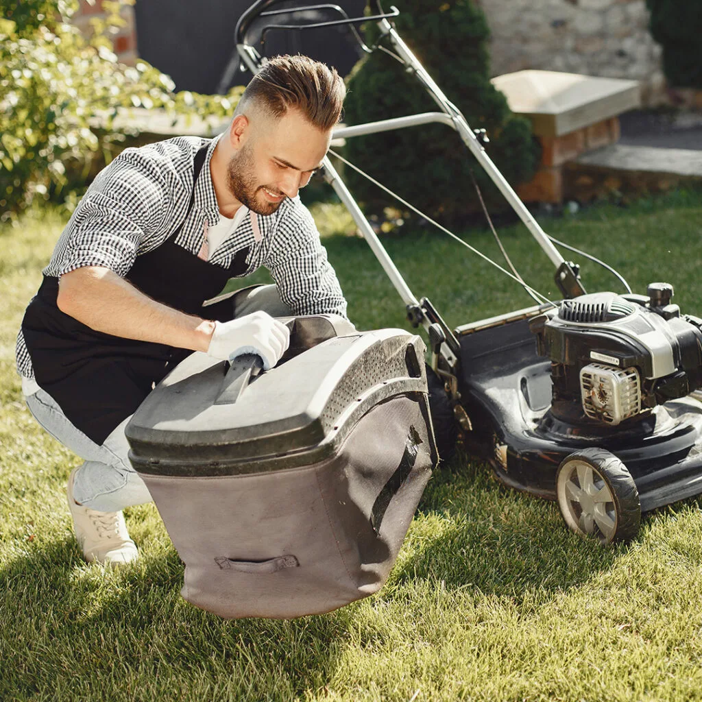 Man in apron and gloves attaching a grass catcher to a lawn mower while performing affordable landscaping near me services.