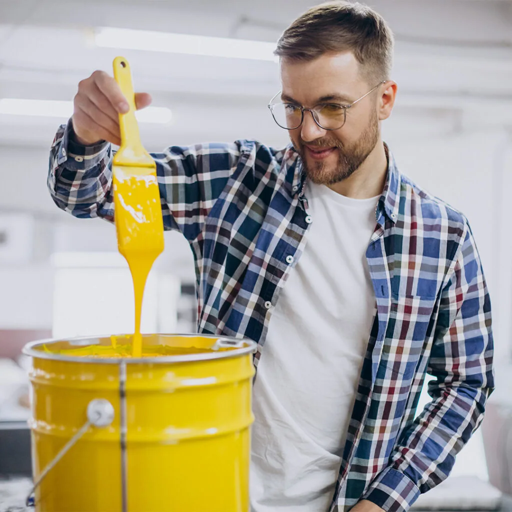 Man dipping a paintbrush into a large yellow paint bucket, preparing for work by professional house painters near me.