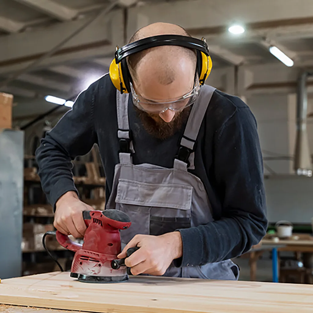 Carpenter near me sanding a wooden surface in a workshop while wearing safety gear and using a handheld power sander.