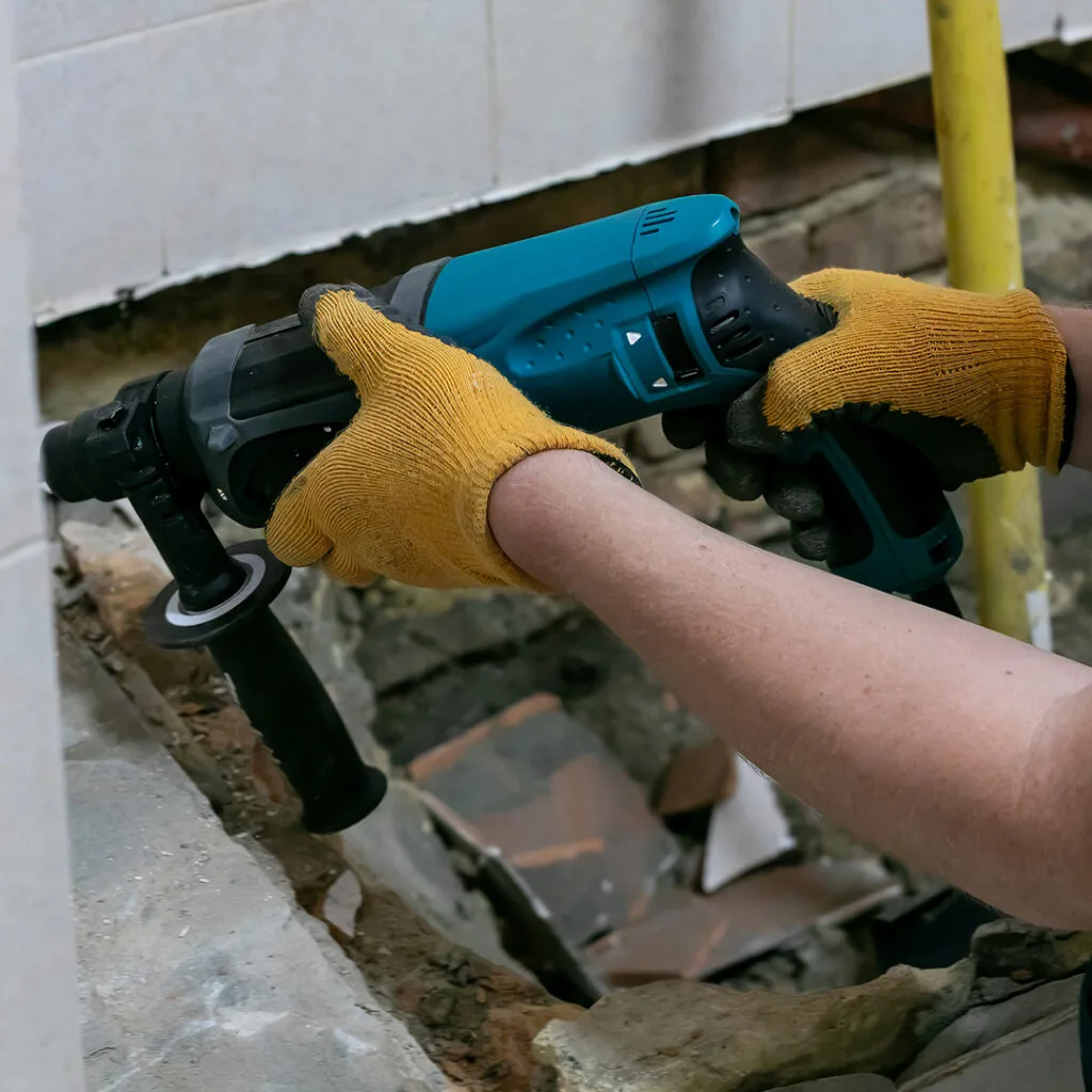 Worker using a power drill to remove damaged tile and drywall during mold remediation near me in a water-damaged bathroom.