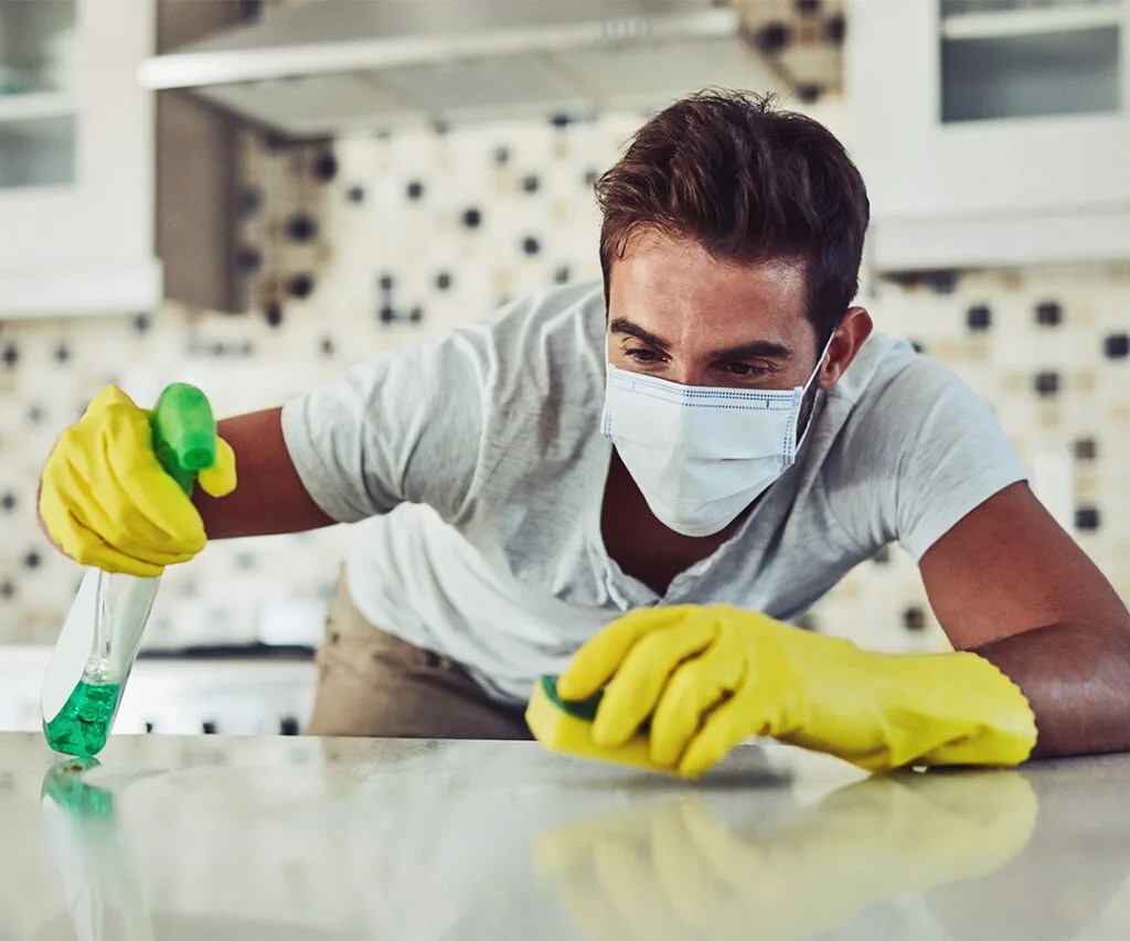 Man wearing gloves and a mask scrubbing a kitchen countertop with spray cleaner, representing house cleaning services near me.