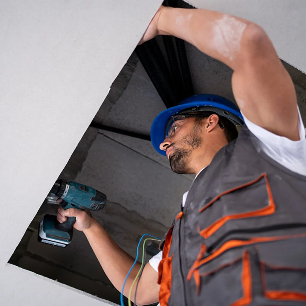 Contractor in a hard hat working on ceiling wiring through a drywall opening during drywall repair near me service.