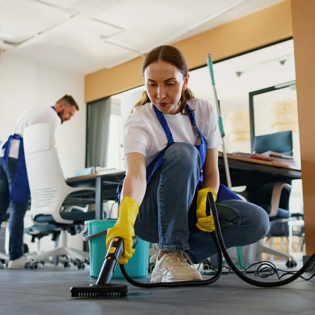 Woman using a steam cleaner on an office floor while a man tidies in the background, representing house cleaning services near me.
