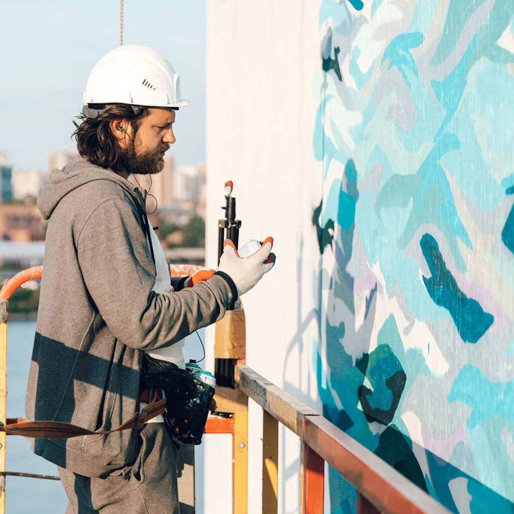 Painter on scaffolding using spray paint to create a mural on a building exterior, showcasing services by house painters near me.