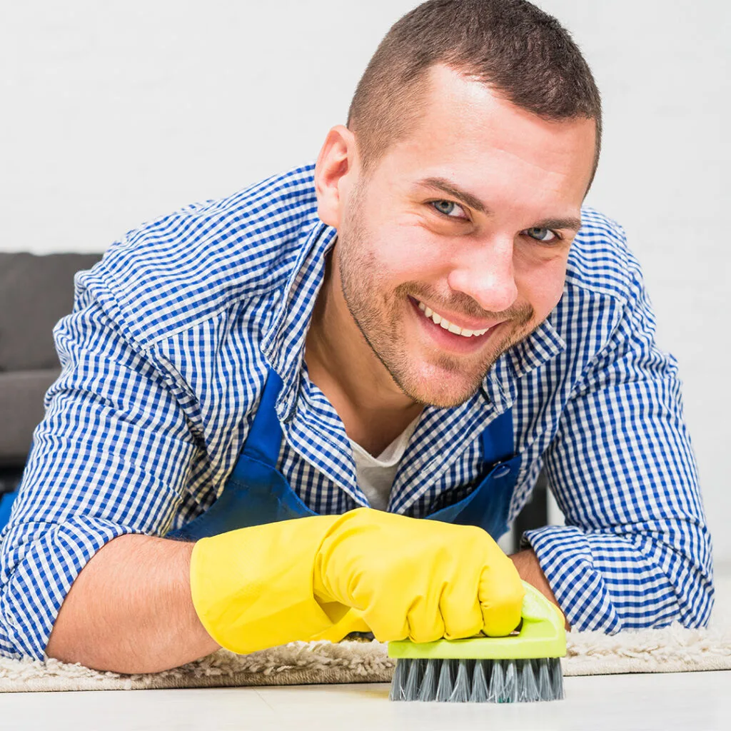 Smiling technician scrubbing a carpet by hand with a brush during a detailed carpet cleaning near me service.