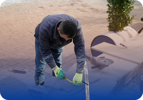 Construction worker measuring a stone slab with a tape measure at an outdoor masonry site.
