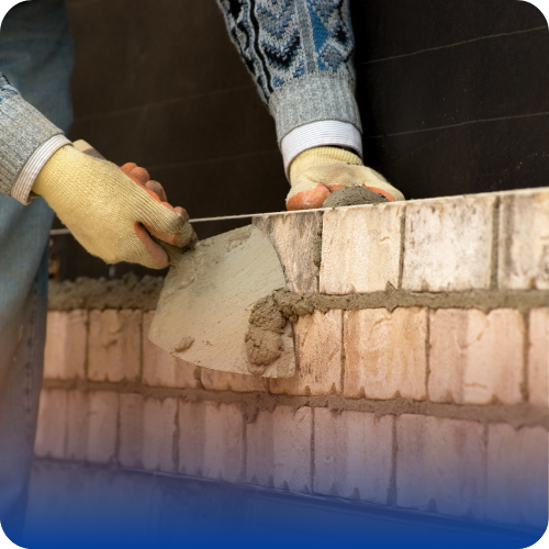 A construction worker wearing gloves spreads mortar with a trowel while laying bricks on a wall