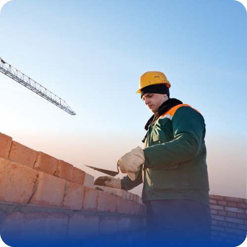 Construction worker in a hard hat and gloves laying bricks and applying mortar on a brick wall at a building site.