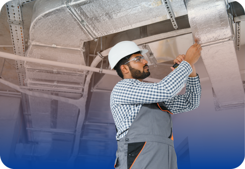 HVAC technician wearing a hard hat and safety glasses inspecting and adjusting ductwork on a ceiling.