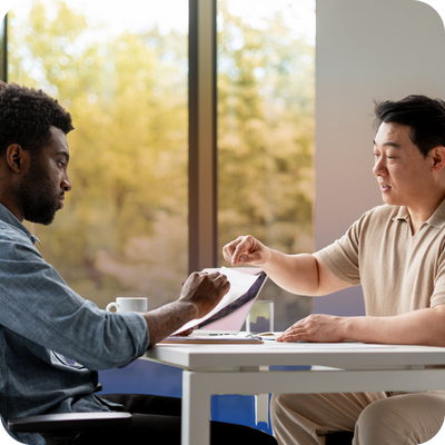 Two men sit across from each other at a table, discussing a document in a bright office with a window view.