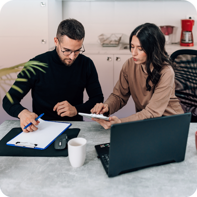 Two professionals collaborate at a table, reviewing documents and notes beside an open laptop in a modern office kitchen setting.