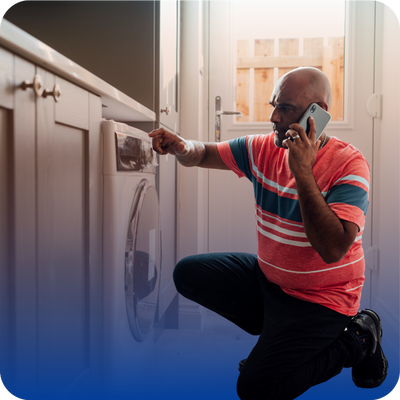 A man kneels in front of a washing machine, inspecting it while talking on a mobile phone in a laundry room—perhaps discussing appliance repair cost with a local pro appliance repair service.