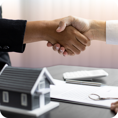 Two people shaking hands over a table with a house model, symbolizing partnership in home contracting and insurance coverage.